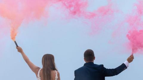 Couple in suit and wedding dress with their backs to the camera - they are holding smoke flares with coloured smoke billowing out.