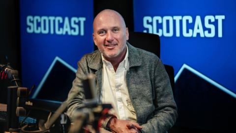 Martin Geissler sits in radio studio in front of blue Scotcast logo