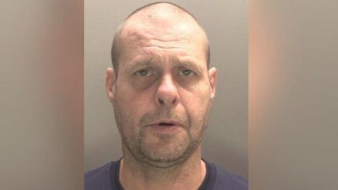 A mugshot of Phillip Colbourne, who is wearing a blue t-shirt and is balding. He has a vacant expression on his face.