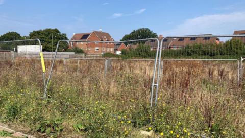 A metal fence in an overgrown area of grassland while in the background homes can be seen.