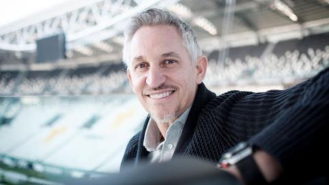 Gary Lineker sits in a football stand smiling, with another stand across the pitch seen behind him out of focus.