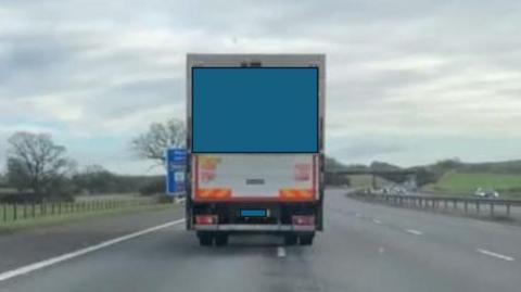 A lorry pictured from the back. The Heavy Good Vehicle (HGV) is partly across the white lines on the motorway. A central reservation and a bridge and railings can be seen in the background. The lorry's markings and number plate have been blocked out by police. 