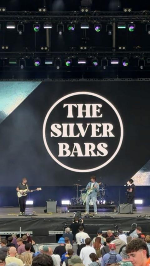 The Silver Bars perform on the main stage at Kendal Calling in front of a large crowd. A giant screen behind the band displays The Silver Bars logo in bold white lettering. Audience members watch from the front of the stage as the band plays under the festival lighting.