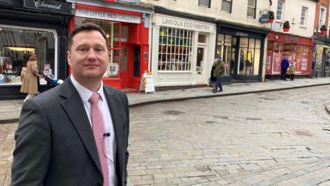 A man wearing a dark-grey suit, white shirt and red tie smiles as he stands on a cobbled street with a row of smart shops, their fronts in various colours, behind him. 
