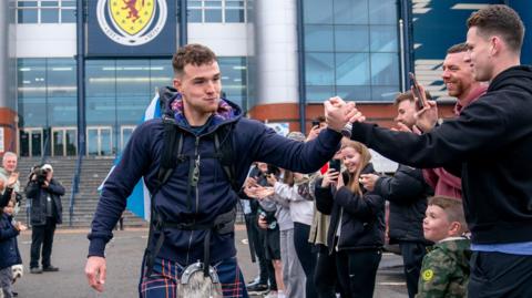 Craig Ferguson wearing a blue red and white kilt and blue hooded top leaving Hampden in the background. He is shaking hands with a man wearing a dark-coloured top. On either side people are applauding.