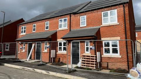 A newly built terrace housing block with orange-brown bricks and white framed windows, with fencing in front of them and a wooden pallet resting next to a front door