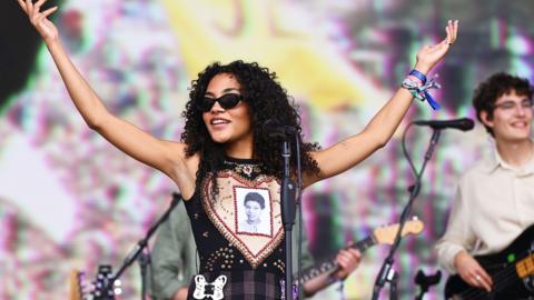 Olivia Dean performing at Glastonbury, wearing a dark, sleeveless shirt and sunglasses, and holding her hands in the air