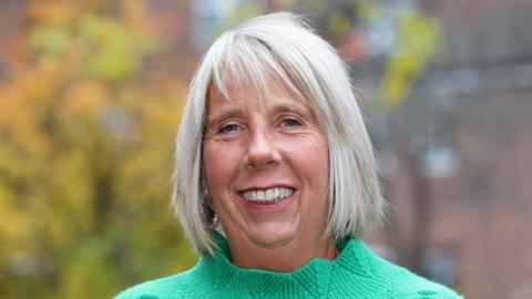 Alison Tatum in a green jumper standing outside with blurred trees and buildings in the background.