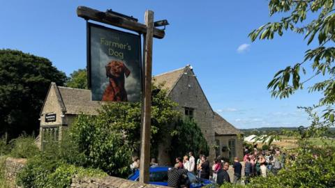 Outside the a village pub. There's a dog on the sign and people queuing outside.