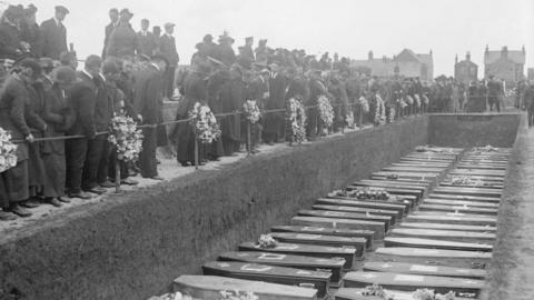 A black and white image of a large grave with dozens of coffins inside. There is a large group of people standing around a rope on the edge of the pit.