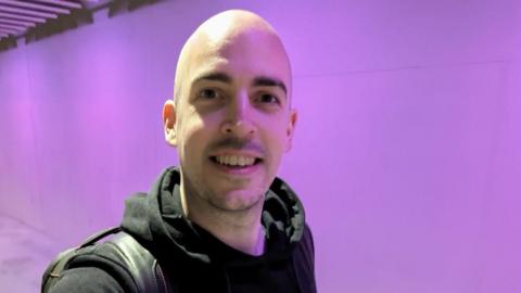 Dean Stokes - a man with no hair smiles into the camera. He has a black hoodie on and has a black bag over his shoulder.