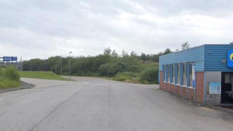 A road into an industrial estate. A blue clad car wash on the right. A grassy bank on the left and fence in the distance with a blue sign on it.