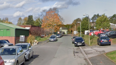 A street with cars parked on both sides with some grassy verges. Industrial-looking buildings are on both sides of the street.