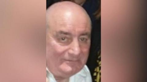 A older man looking at the camera. He is bald and wearing a white shirt