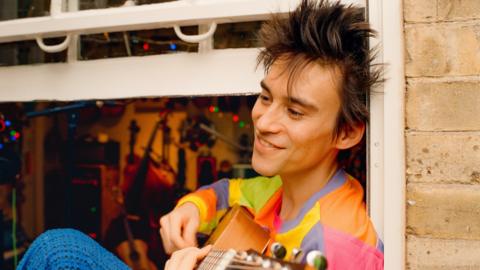Jacob Collier sits in an open window holding a guitar. He's a man in his thirties with spiky long brown hair. He's smiling and wearing a multi-coloured patchwork t-shirt and blue trousers. Behind him musical instruments are visible through the open window. 