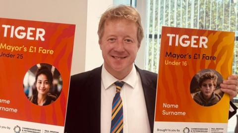 Paul Bristow is wearing a black suit jacket, white shirt and colourful striped tie while holding two orange placards. They advertise the Tiger scheme and its £1 fares.