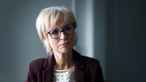 Sally Magnusson sits in a darkened room wearing blue-rimmed glasses, a white top and a burgundy blazer