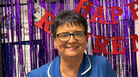 A woman in a blue nurse uniform and glasses is smiling at the camera in front of purple string tinsel and red lettering which reads ' Happy Retirement' 