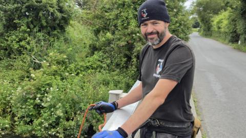 Gareth Morris stands on a bridge holding a rope before he begins magnet fishing. He wears a black tee-shirt and jeans and a black beanie hat with a magnet on it. 