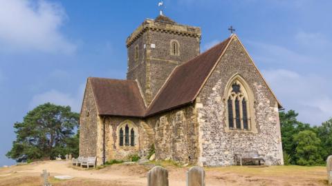 A small grey stone church on a hill with gravestones in the foreground and green trees in the background