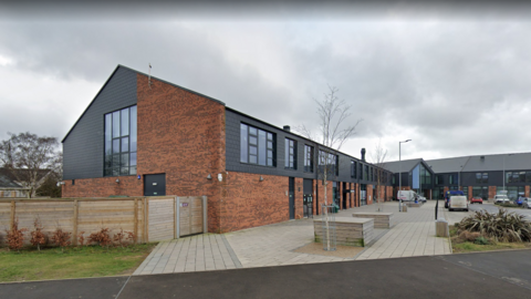A school building made of brick and dark wood panelling with a paved area in front, wooden fencing and small grassy areas. There are cars and vans parked outside.
