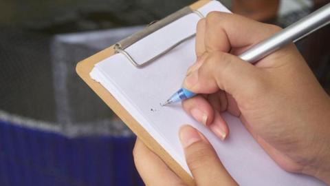 An image of a woman with a pen in her right hand, writing on a clipboard.