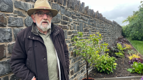 John Watkeys standing against his new stone garden wall which is about head height. He is wearing a fawn-coloured hat and glasses with a black coat and has a white beard. A flower bed can be seen to the right of the wall and grass to the right of that. 