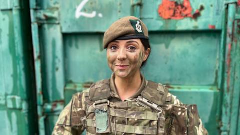 Rakela Kalemi wearing army fatigues, an Army beret and camouflage paint. She is standing in front of a copper green shipping container