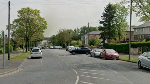 The image shows a suburban street on a clear and sunny day. Cars are parked along both sides of the road. Trees line the street and houses are visible in the background, suggesting a residential area. Power lines and poles run alongside the road.