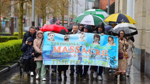 People walking carrying a large banner reading: "Springhill-Whiterock Massacre Belfast's Bloody Sunday Time for Truth" with pictures of the five deceased. The people are all wearing raincoats and carrying umbrellas in the rain.
