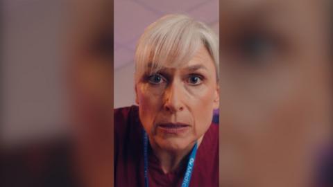 Close up of woman with a grey fringe and purple scrubs, who peers at the camera.