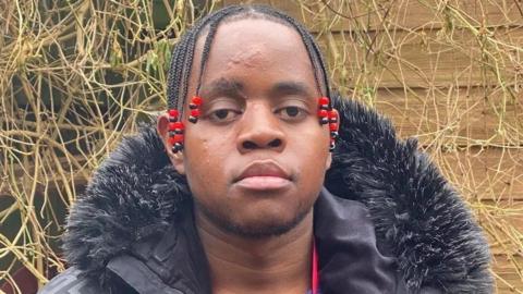 Katungua Tjitendero. He has short dark hair which is in several little braids. They have red beads on the ends. He is pictured looking directly at the camera with a blank expression. He is wearing a black coat with a fur lined hood. 