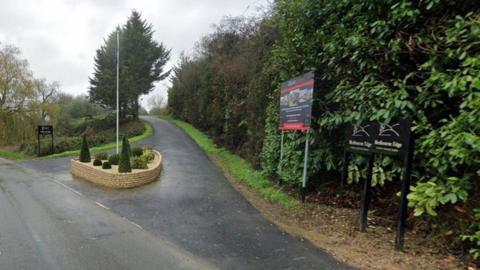 The entrance to a holiday lodge site on a country lane, with a nice piece of planting and a flag pole in an island at the entrance, and some signage around the edges