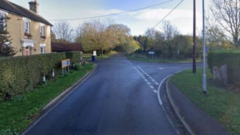 A view of the junction from Hilton Road into Connington. There is a house on the left side, behind a hedge, and a road sign to Connington and Fenstanton next to the junction. There are telegraph wires overhead and two posts on the right. Trees and fields are in the distance.