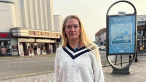 Lisa Pearson with long blonde hair wearing a white cricket-style sweater, with black bands around the neck. She is looking at the camera. "The Old Savoy", an art deco building, is visible in the background on the other side of a road. A blue advertising unit is showing a "Be More Us" poster.