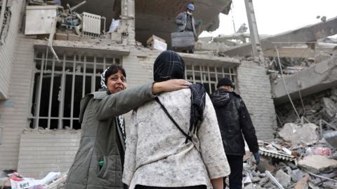 A woman holds the shoulder of another woman in front of a collapsed building with rubble all around in Tehran on 12 March.
