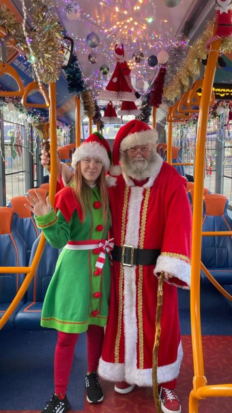 Frances is dressed as an elf in a green dress and wearing a Santa hat, standing next to her dad Thomas, who is dressed as Father Christmas in a red and white long coat. They are stood on the bus with lots of fairy lights and decorations around them.