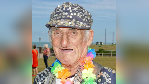 An elderly man in a blazer and cap adorned with buttons. He is wearing a ring of flowers around his neck. 