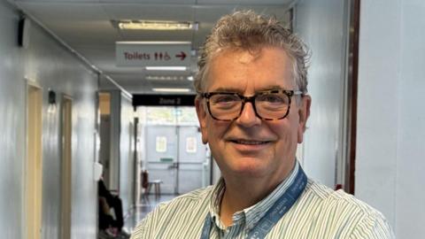 Dr Thomson is standing in a hospital corridor, wearing glasses and a white shirt with pale blue and grey stripes. He is smiling and has a lanyard around his neck.