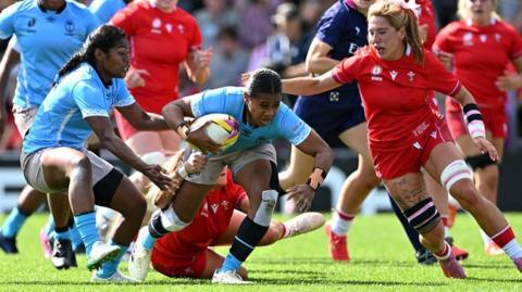 Josivini Naihamu in blue jersey is tackled as Georgia Evans wearing a red jersey and red shorts moves in