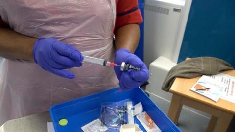 A nurse wearing blue gloves holds a syringe in their hands above a blue tray with medical equipment.
