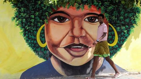 A boy walks past a wall with a woman's face painted on it. She has a green afro which blends into the trees growing behind the wall, giving the illusion that the trees are part of the artwork.