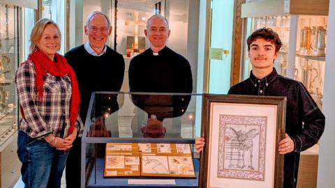 Charlie Norman stands holding his artwork in St Edmundsbury Cathedral alongside Louise Griddle and members of the cathedral. Mr Norman wears a dark shirt and has dark hair.