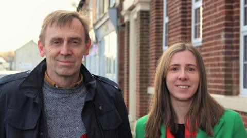 Katie Weston and Paul Weston are standing side by side in front of a red brick house. They are both in their 40s, she has shoulder-length brown hair and is wearing a green jacket, he is a little older with a blue jacket and light blue jumper. They are both smiling.