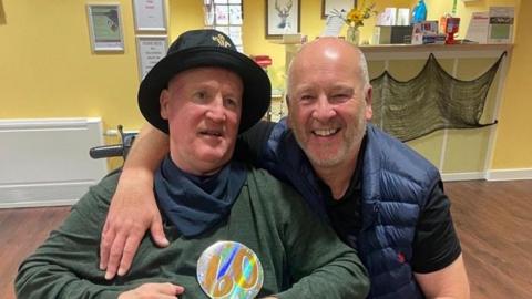 Anthony has grey hair and beard in a blue gilet with a black t-shirt underneath. He has his arm round his brother Keith who is in a wheelchair and is wearing a green long-sleeved t-shirt with a navy bandana round his neck and a black hat on.