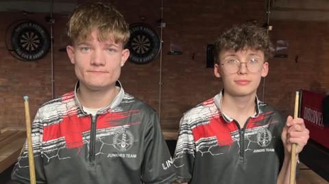 Jed Leech and Cavan Roberts standing in front of a pool table, with dart boards behind them. They're holding their cues and wearing their England team zipped tops. 
