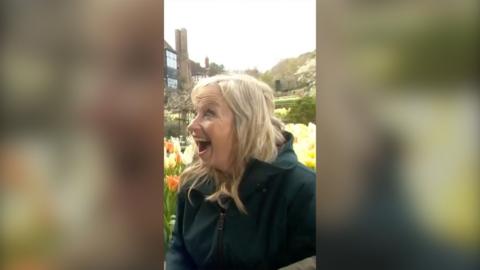 Carol Kirkwood wears a green coat and smiles in surprise against a backdrop of yellow and orange flowers