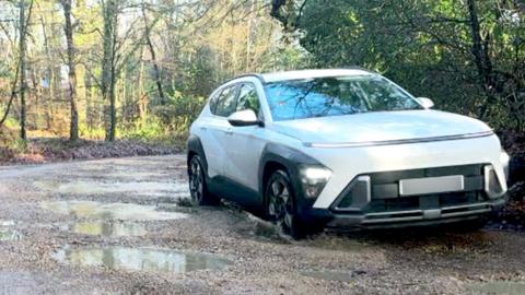 A white car driving through pot holes in a New Forest car park