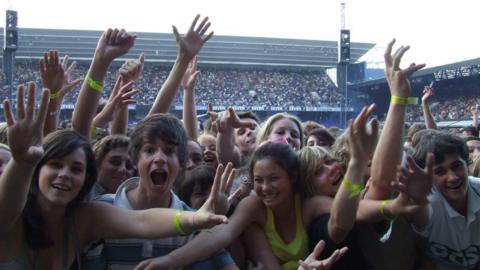 Red Hot Chili Peppers fans smile at the camera with their arms in the air during a performance at Portman Road stadium. Thousands of fans can be seen in the stands behind them. 