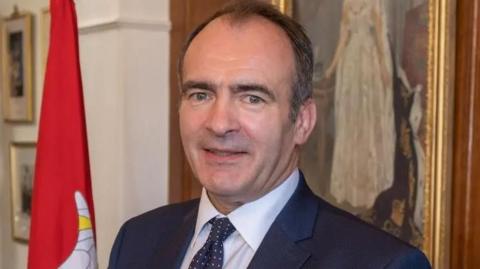 Alfred Cannan smiles into the camera. He is wearing a navy blue suit and tie with a white shirt and he is standing next to a red Manx flag.